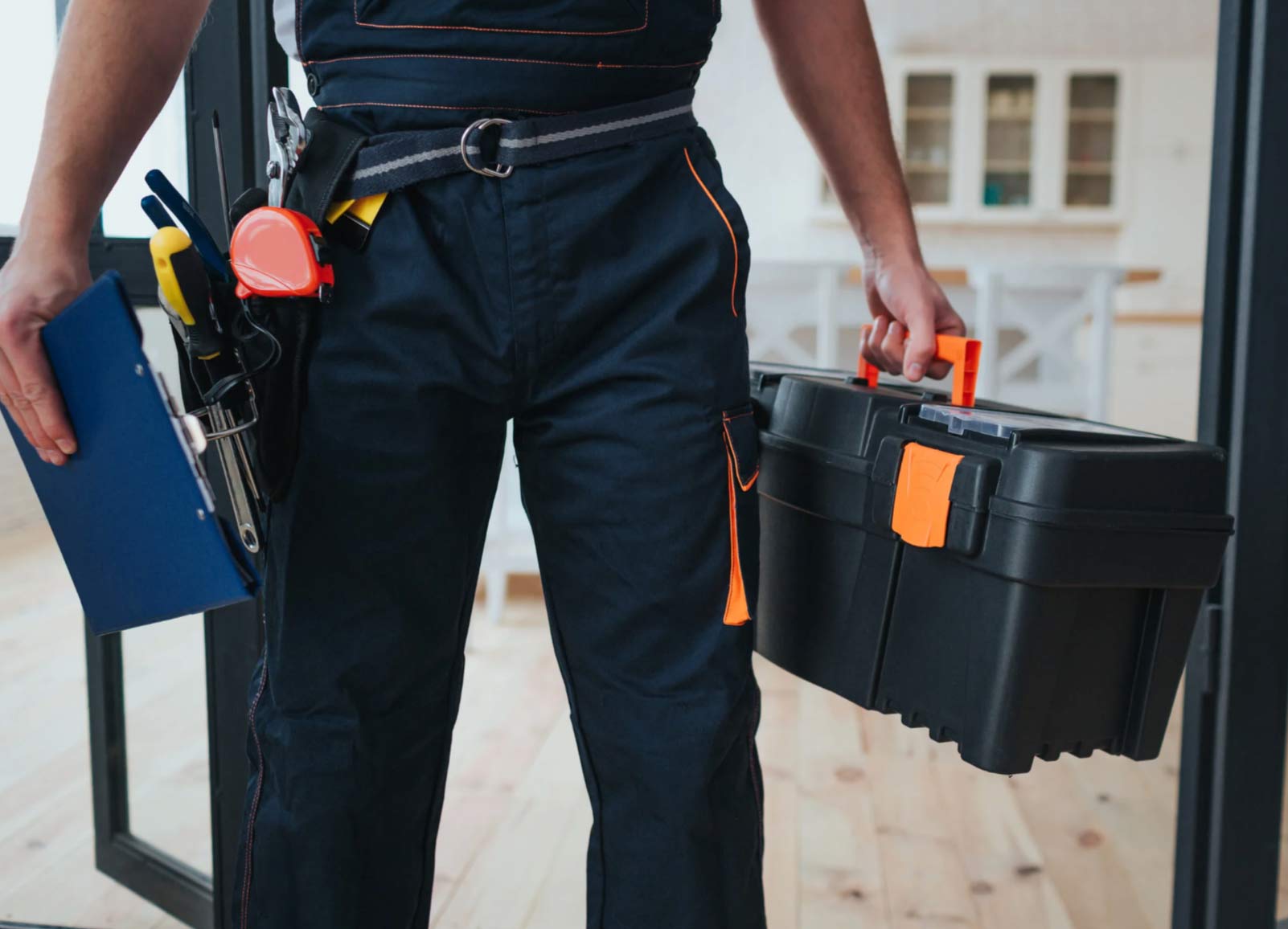 A worker in overalls holds a toolbox and a folder, with tools attached to a belt around his waist.