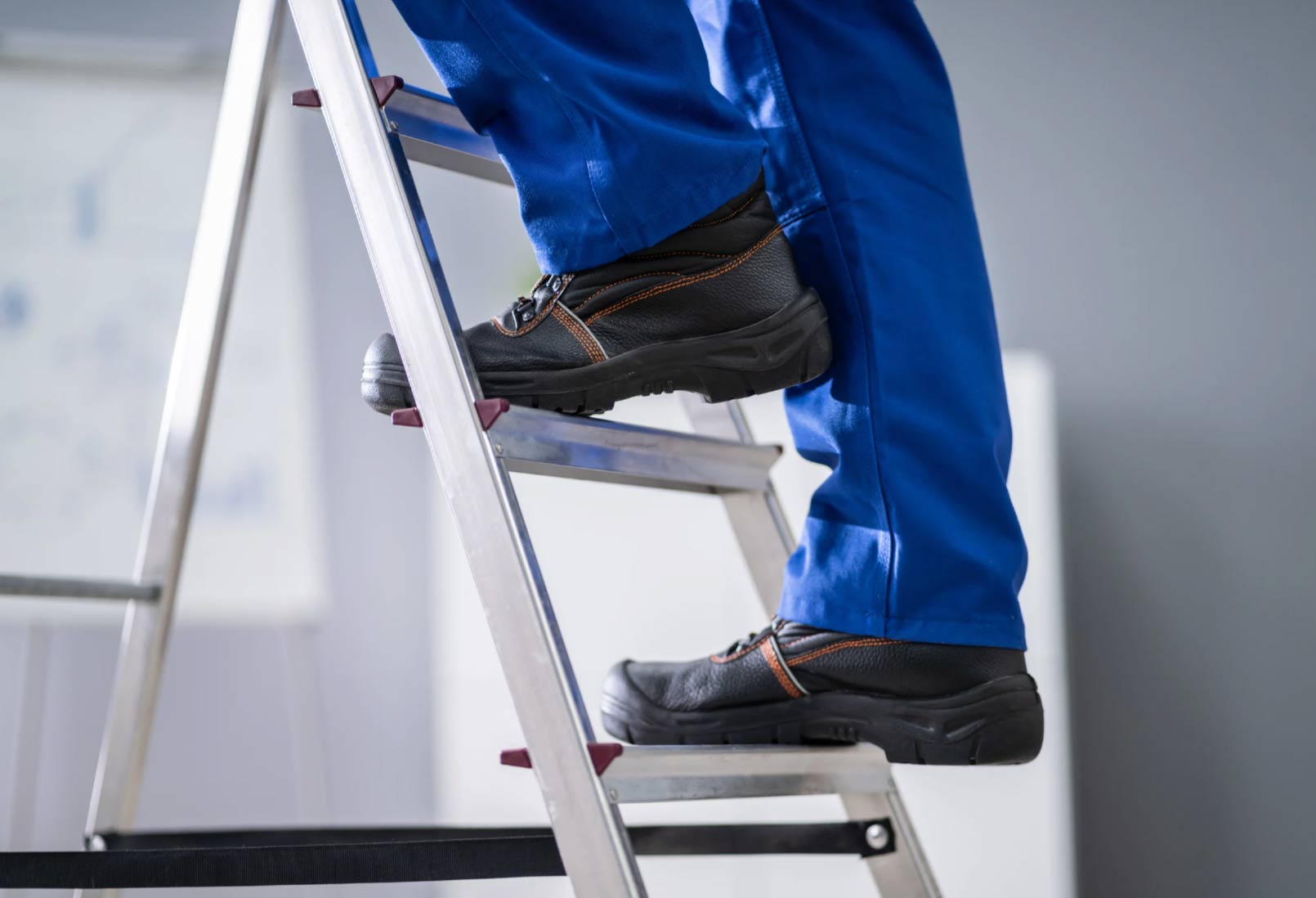Person in blue work pants and black safety shoes standing on a metal ladder indoors.