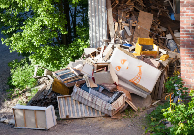 A pile of discarded wood, furniture, and a board with a smiling face near a building and trees.