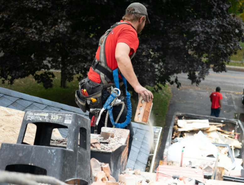 A construction worker in a harness removes debris from a roof and tosses it into a bin below.