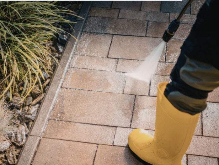 Person in yellow boots pressure washing a tiled outdoor walkway near some plants.