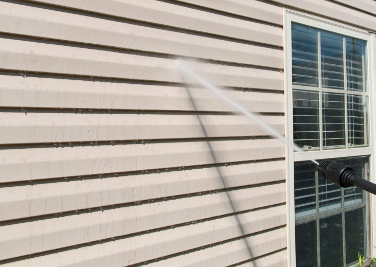 A pressure washer sprays water on the beige siding of a house next to a window with white trim.