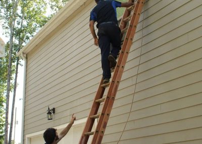 Two people use a tall ladder to reach a high window on the side of a beige house on a sunny day.