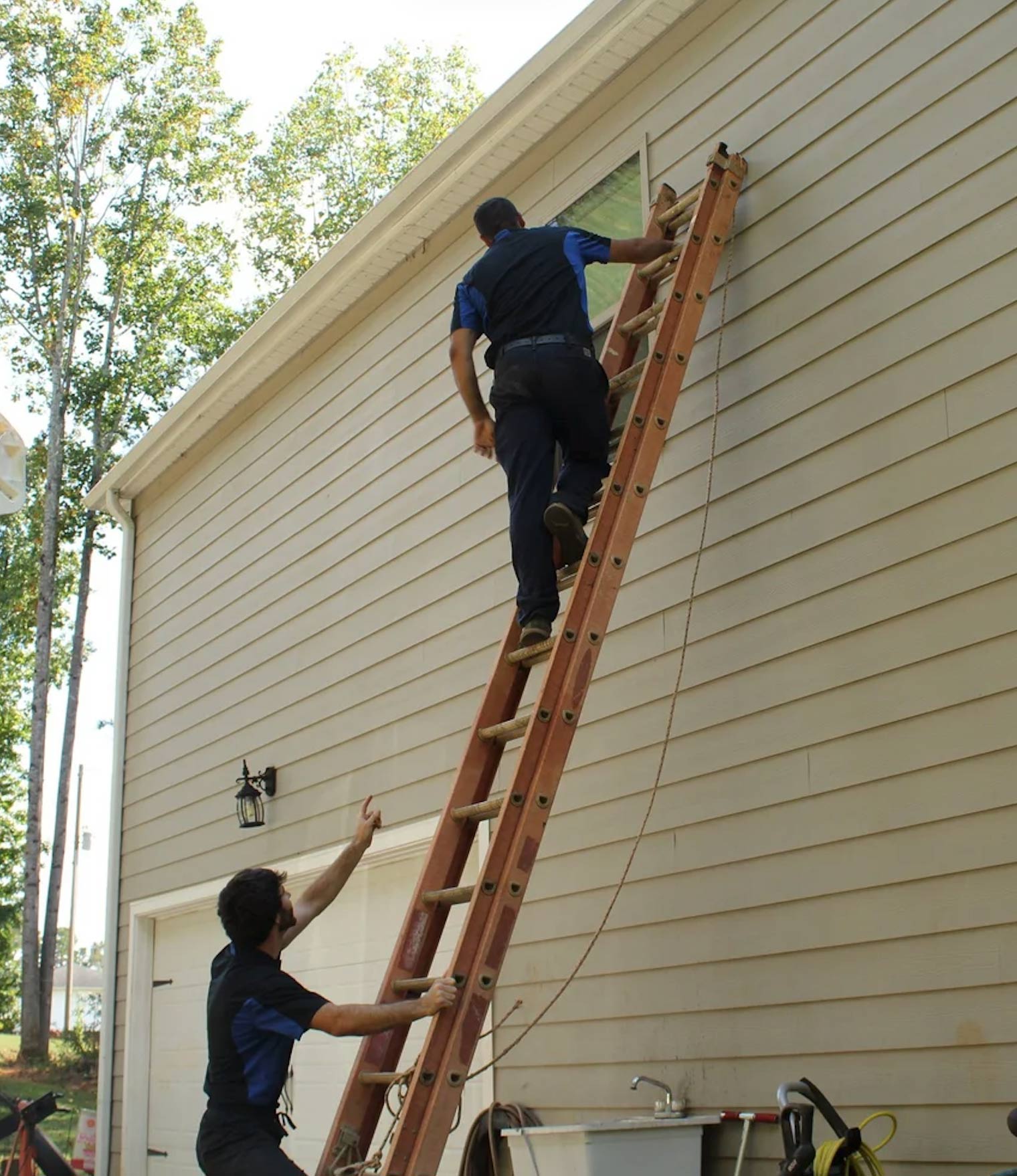 Two men working, one climbing a tall ladder against a house while the other steadies it from below.