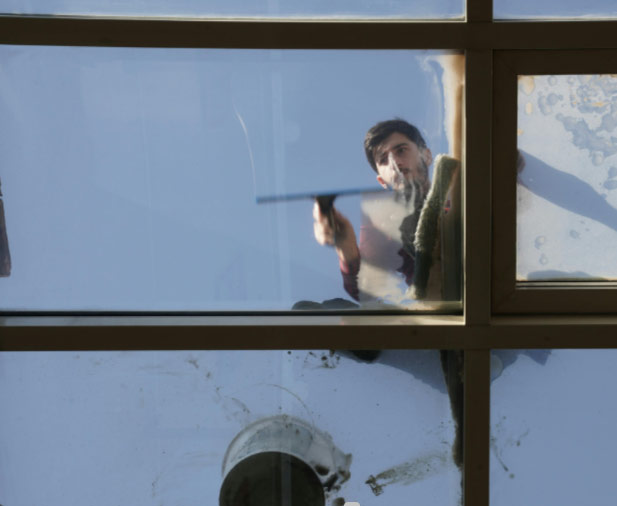 A man seen through a glass ceiling cleans the window with a squeegee against a blue sky.