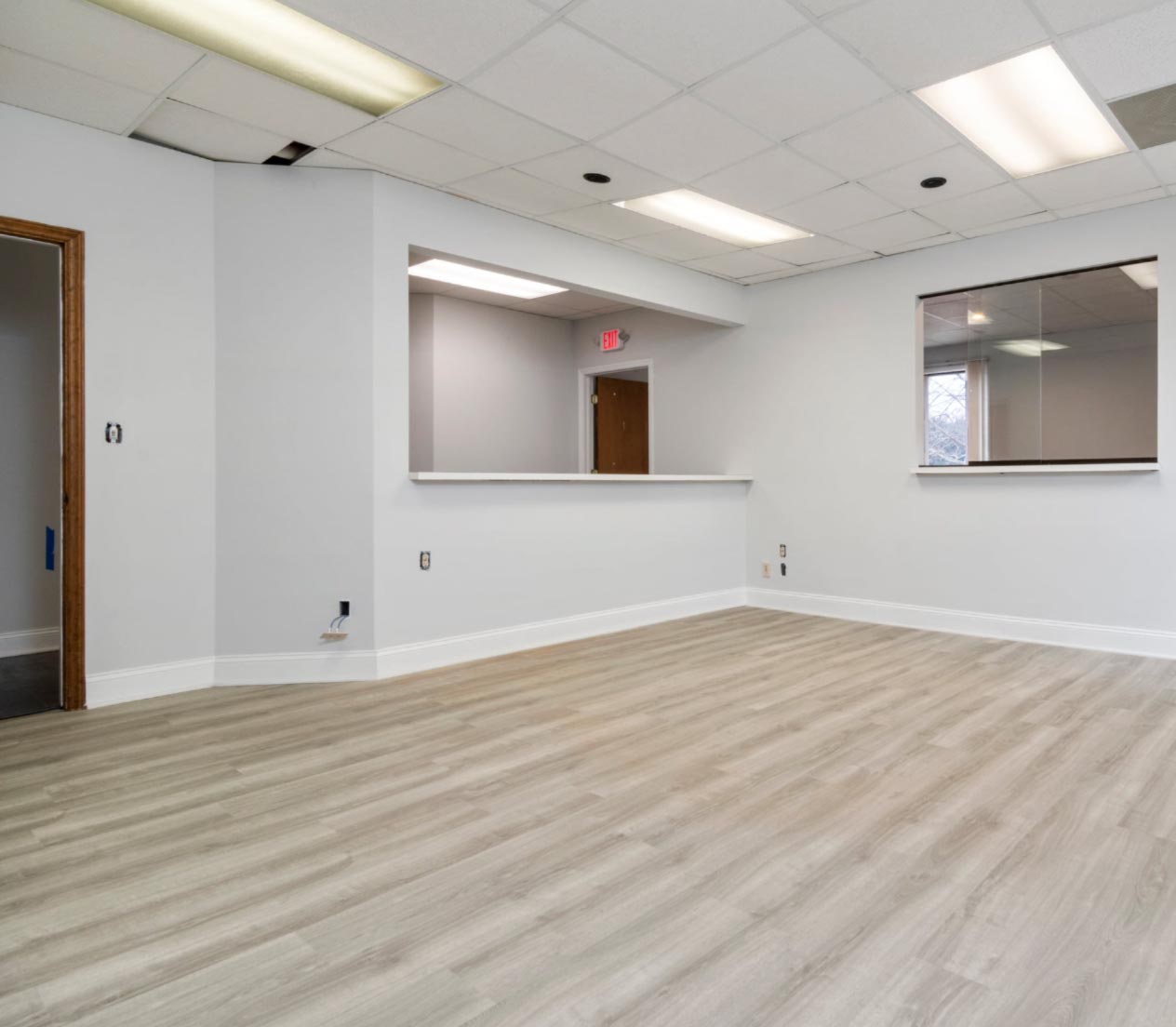 Empty office space with light wood flooring, white walls, ceiling tiles, and a large interior window.