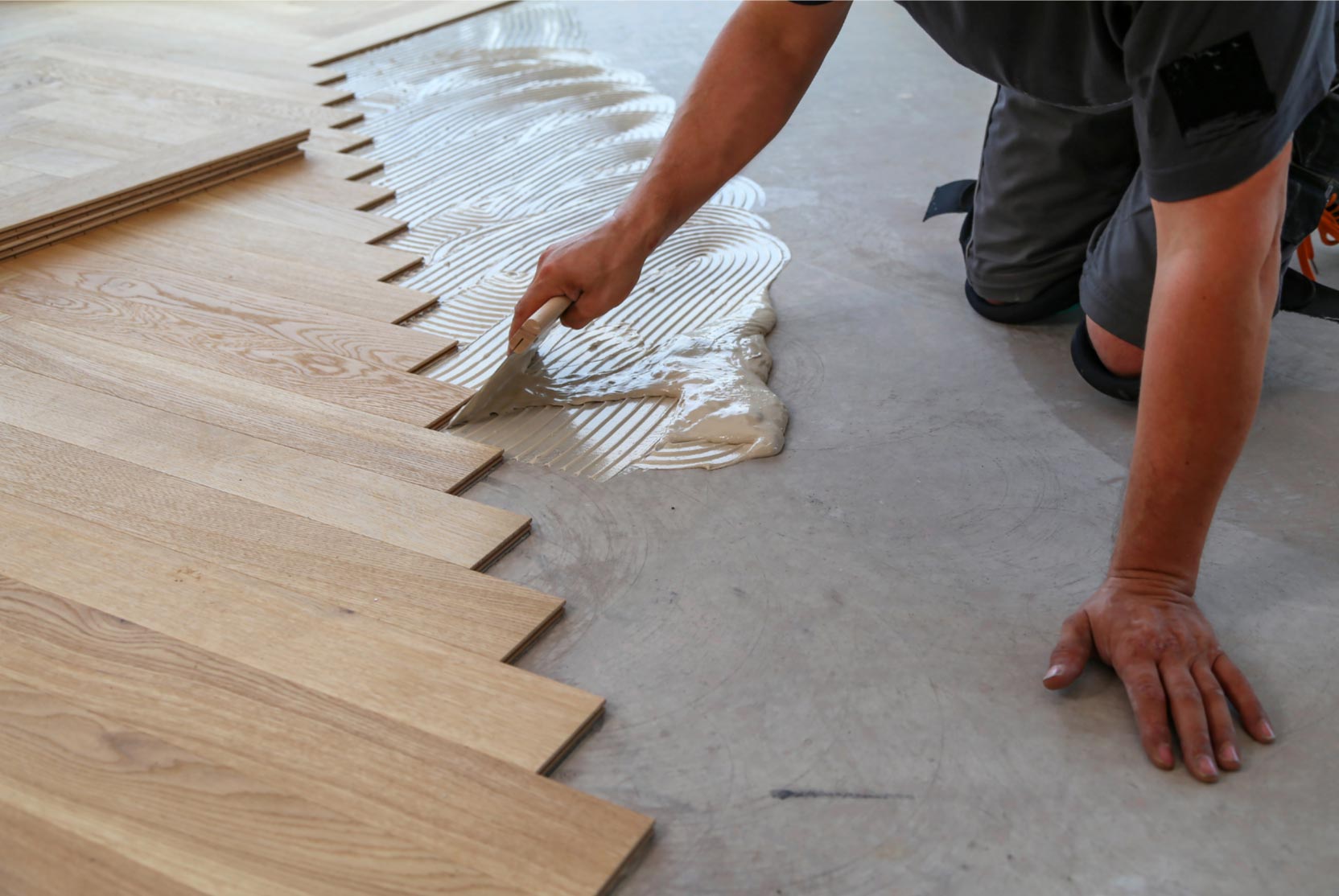 Person installing wooden flooring, spreading adhesive on concrete floor with a trowel, staggered planks visible.